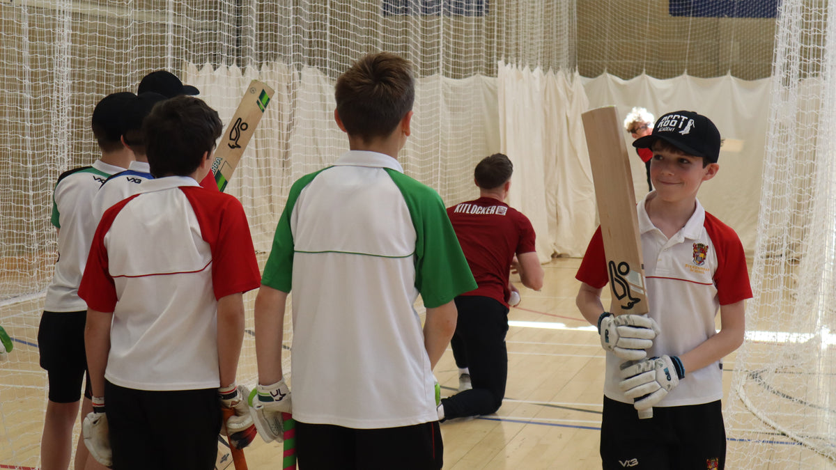 Children in cricket uniforms holding bats and gloves in a indoor cricket facility.