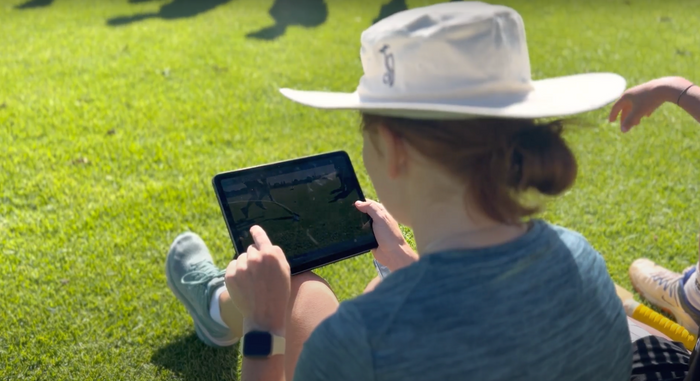 Coach sitting on grass watching back her teams cricket on an Ipad
