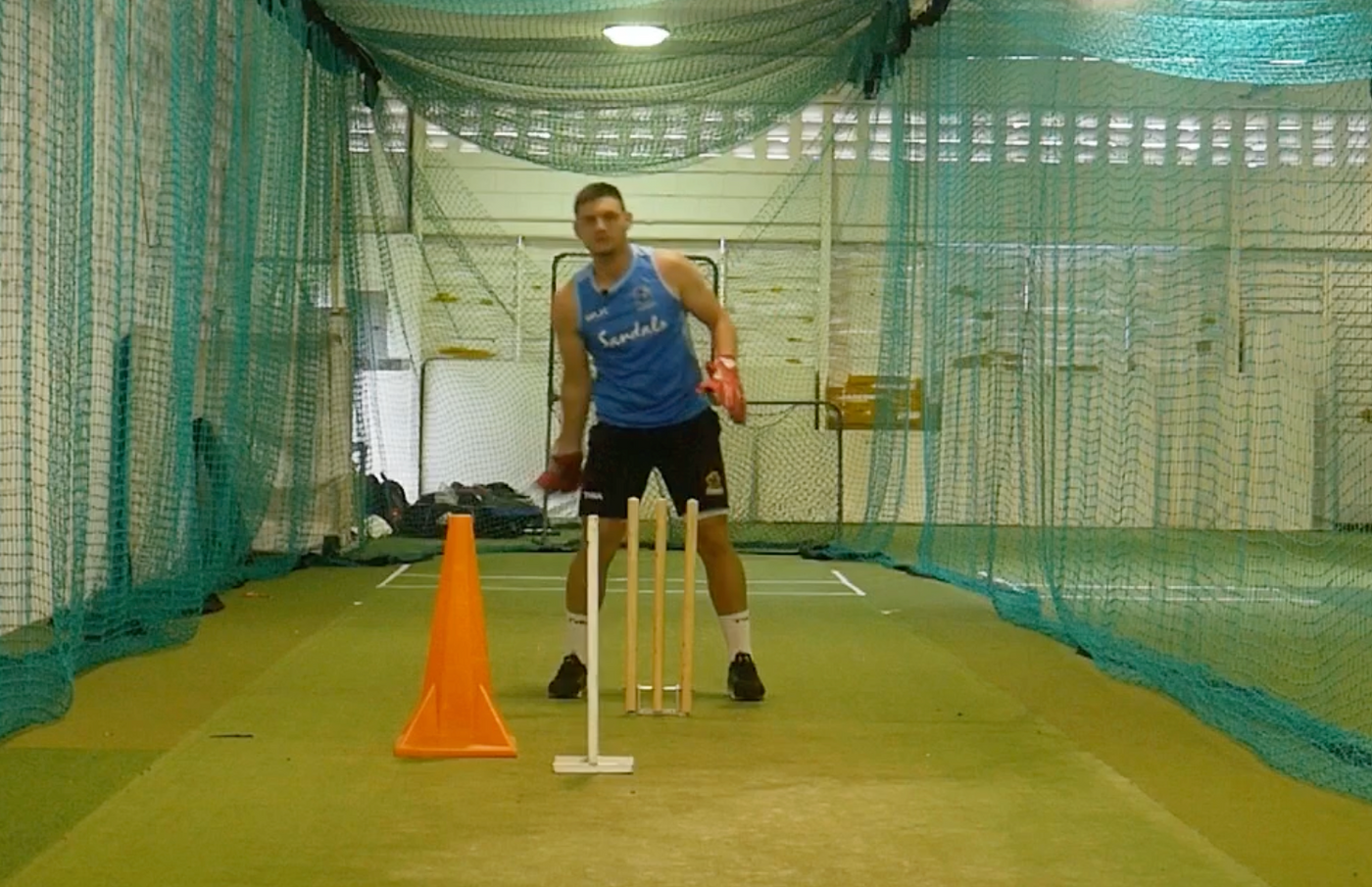 Person practicing cricket in an indoor facility with nets and a cone.