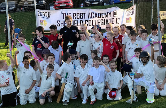 Group of young cricket players and Joe Root posing for a photo in front of a banner at a sports event.