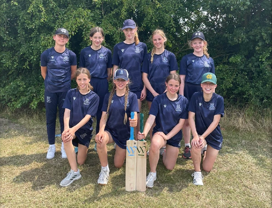 Group of young cricket players in blue uniforms posing outdoors with a bat.