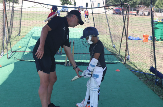 Coach teaching a young player cricket batting techniques in a net session.