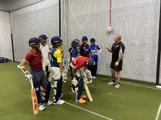 Group of children practicing cricket in a indoor facility with a coach.