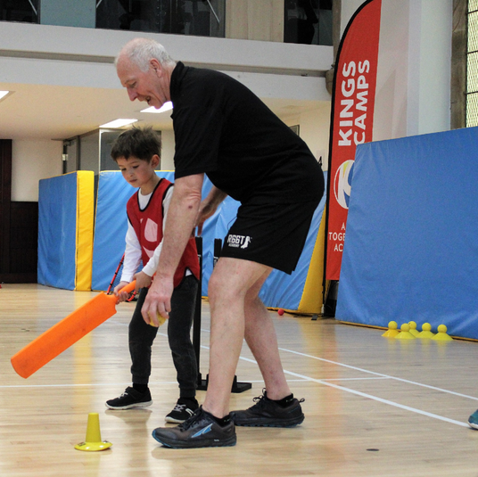 Man and child practicing with a bat on a sports court, with 'Kings Camps' branding in the background.