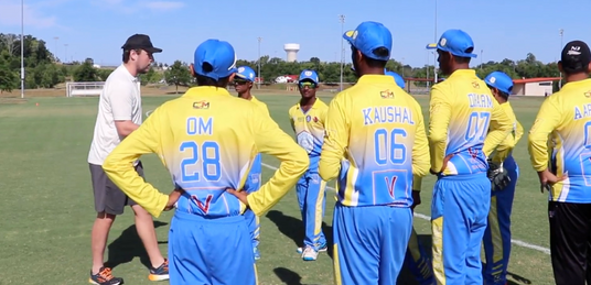 Group of cricket players in blue and yellow uniforms on a field with a coach.