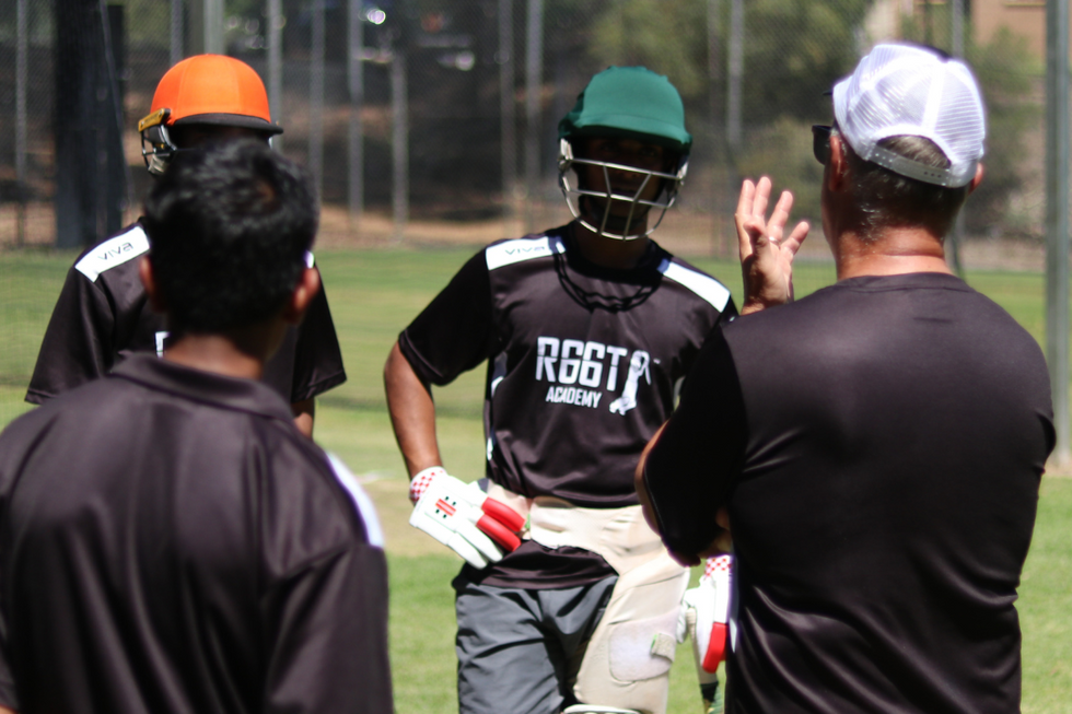 Cricket players and coach on a field with trees and fence in the background