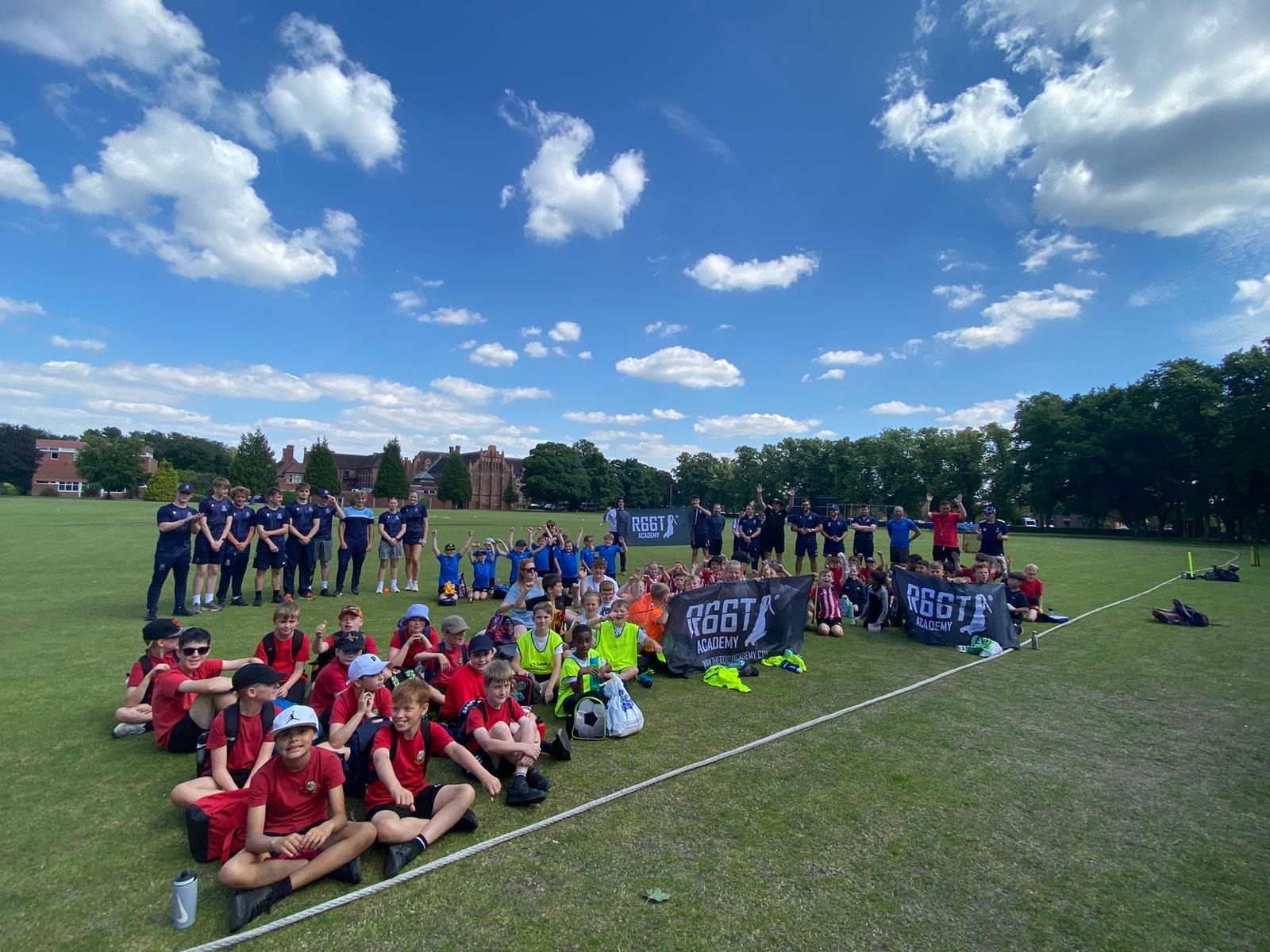 Groups of cricket teams sat outdoors on a grassy field with three 'R66T Academy' logos
