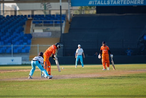 Cricketers in orange and blue uniforms on a cricket field with a blue banner in the background.