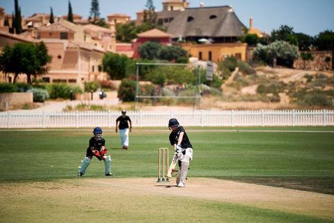 Children playing cricket on a grassy field with buildings in the background