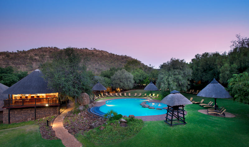 A scenic view of a pool area with thatched-roof structures and a building at dusk.