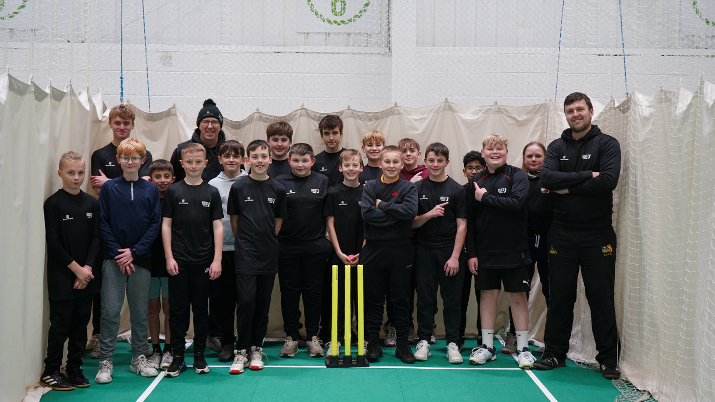 Group of children and adults in a cricket indoor facility holding cricket equipment.