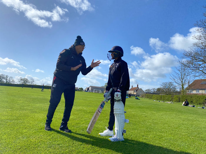 Child in cricket kit being coached on a grassy field.