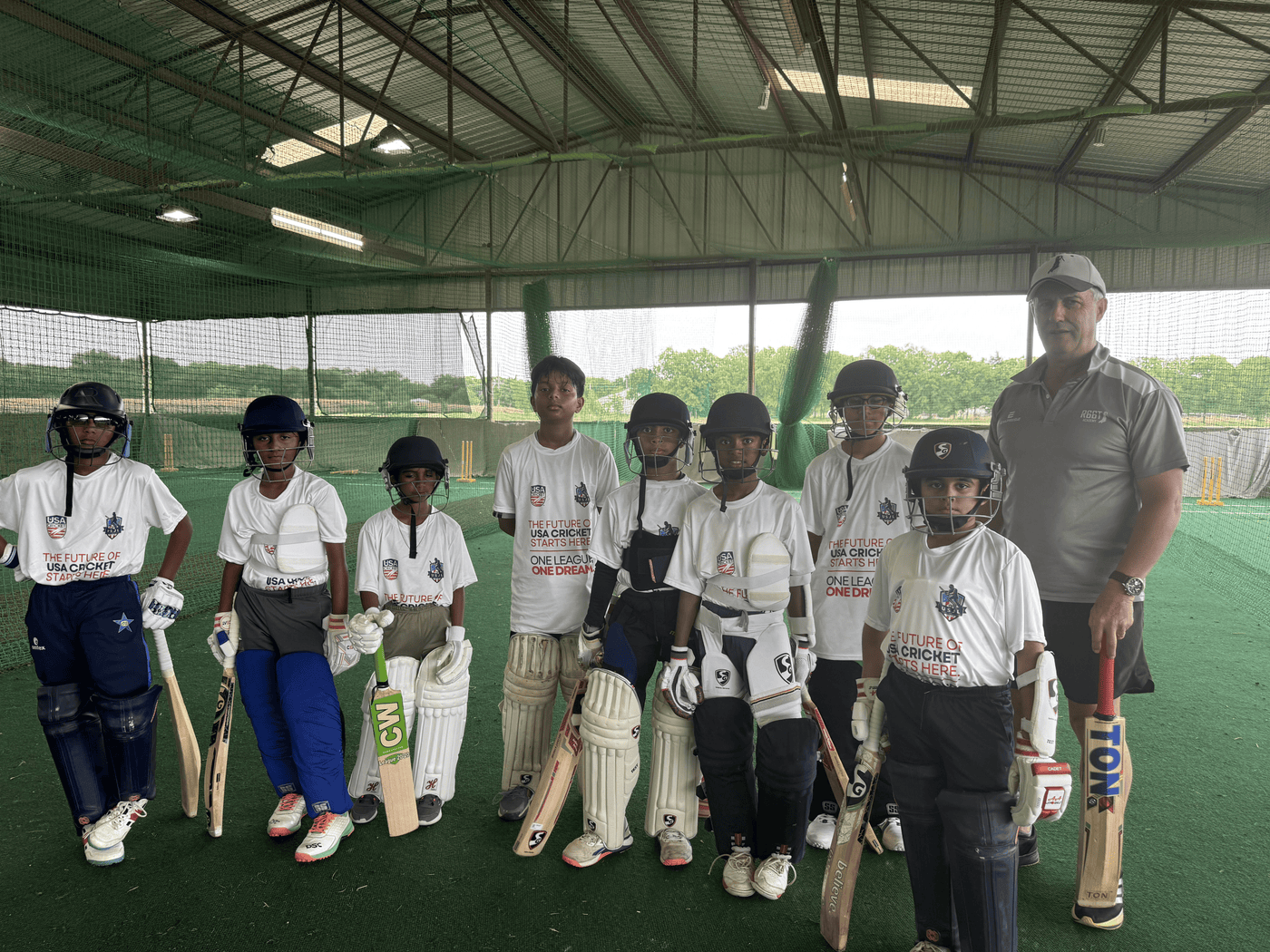 Group of children in cricket uniforms with coaches, holding cricket bats and gear indoors.