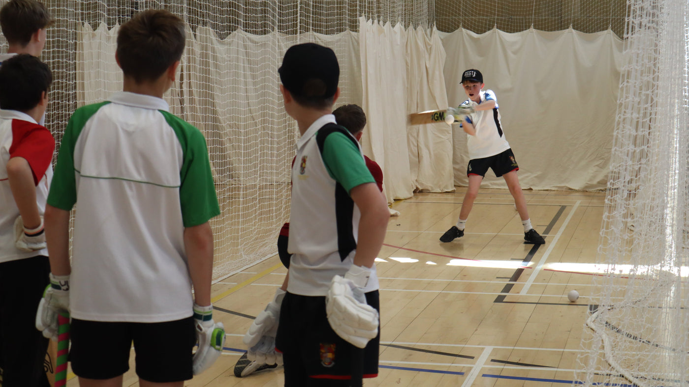 Children playing cricket in an indoor sports facility with a net and white curtains.