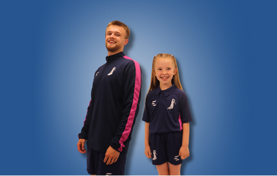 A man and a young girl modeling matching navy blue athletic tracksuits with pink side panels and white logos, set against a solid blue gradient background.