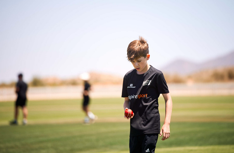 Young boy on a sports field holding a sports ball, wearing a black shirt with 'R66T Academy', 'inspiresport, and 'Emprise' branding.