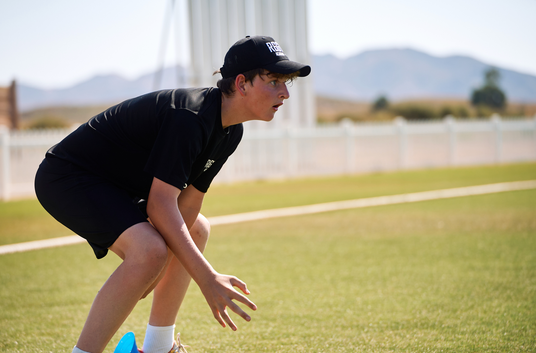 Person in black outfit and cap on a sports field with mountains in the background