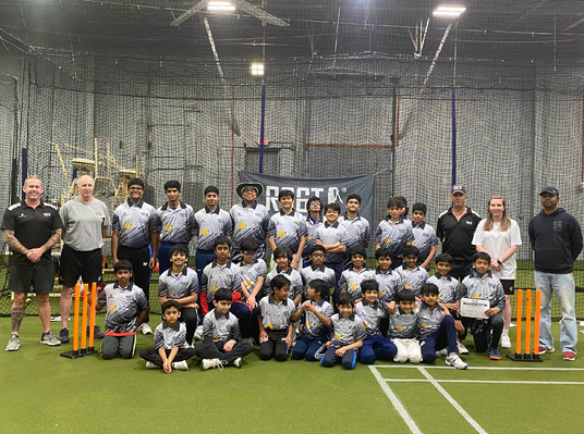Group photo of a cricket team and coaches in an indoor sports facility.