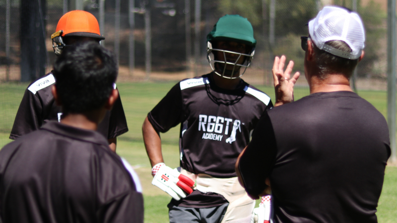 Cricket players and coach on a field with trees and fence in the background