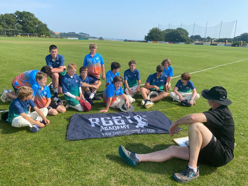 Group of children in sports attire sitting on a grass field with a banner in front.