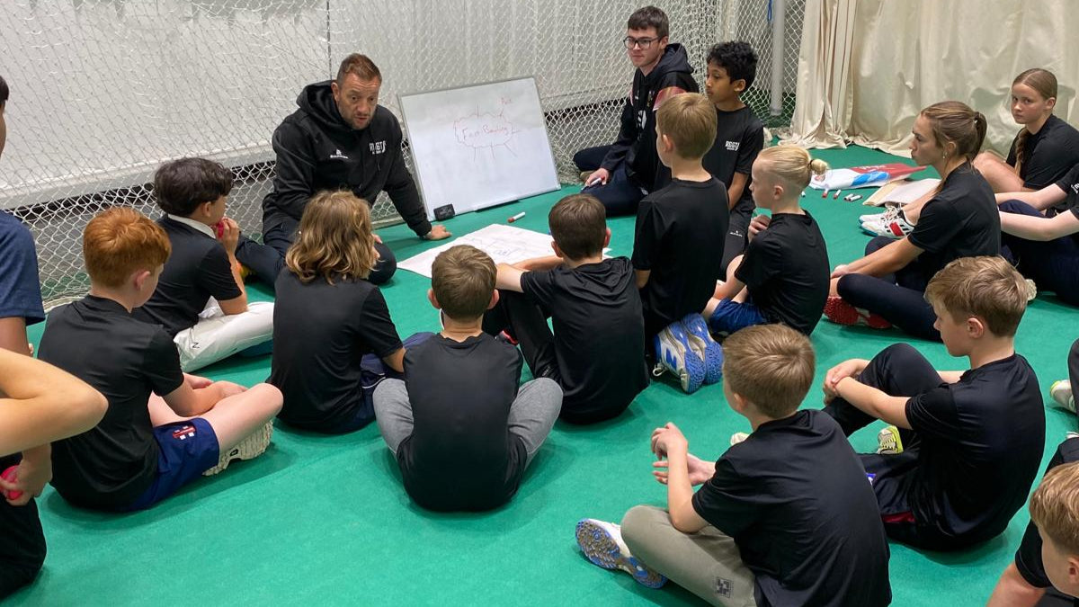 Group of children sitting on the floor in a circle with a teacher or coach, in an indoor sports facility.