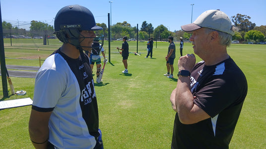Two men on a sports field, one wearing a helmet and sunglasses, the other with a cap and watch, with other people in the background.